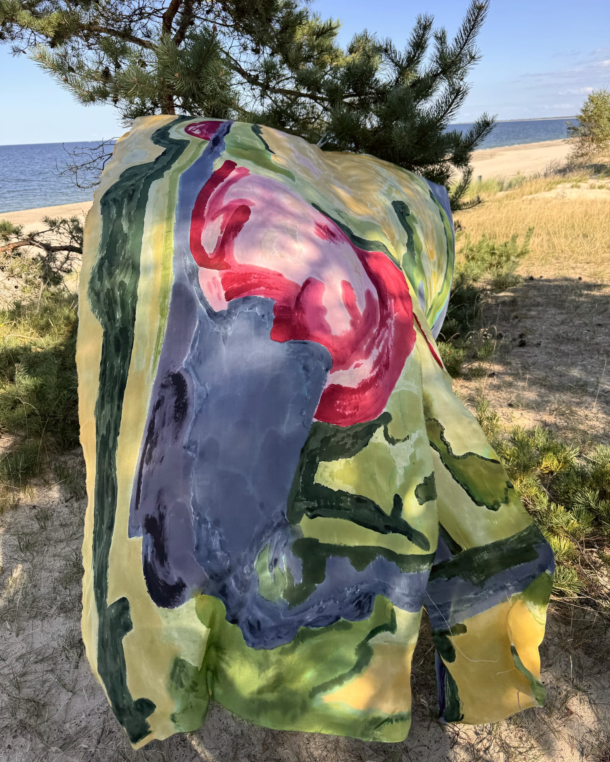 Jedwab w żółcieniach i szarościach z karminowym pąkiem, przewieszony przez sosnę nad plażą.
